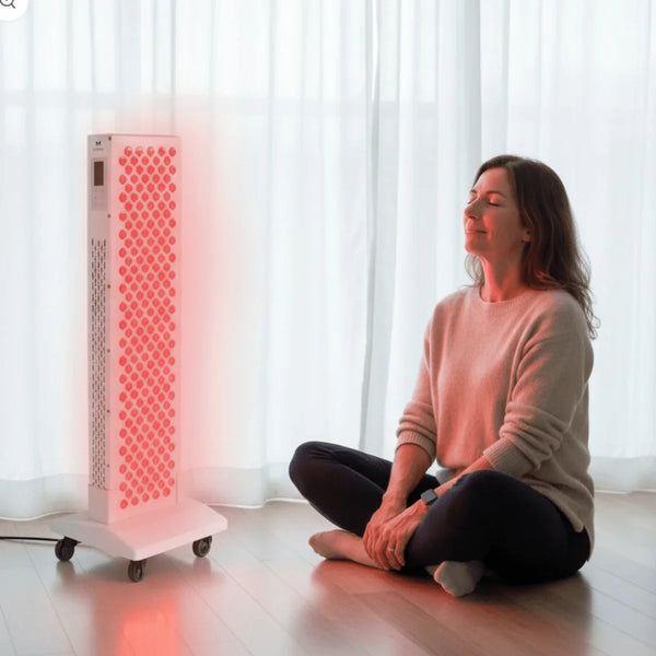 Vrouw zit rustig naast een roodlichtpaneel in een lichte kamer, passend bij een consistente glow routine